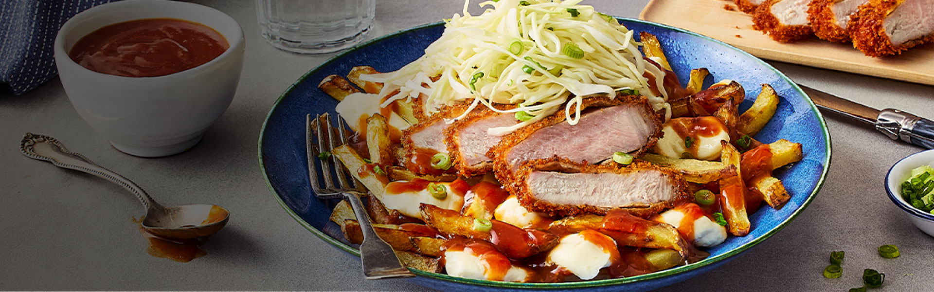 A bowl with the Japanese Tonkatsu Poutine, a cutting board with slices of the pork cutlet, a bowl of green onion, and another bowl of the tonkatsu-style gravy.