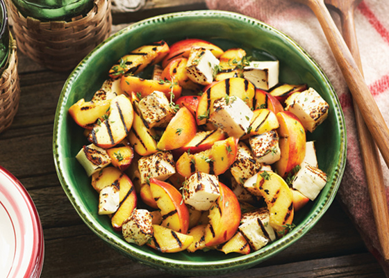 A green bowl filled with grilled peach and haloumi salad on a picnic bench with wooden serving spoons.