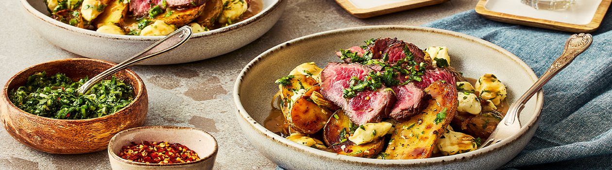 Two bowls of Argentinian poutine with two glasses of lemon water, a bowl of the chimichurri, and another small bowl with red pepper flakes.