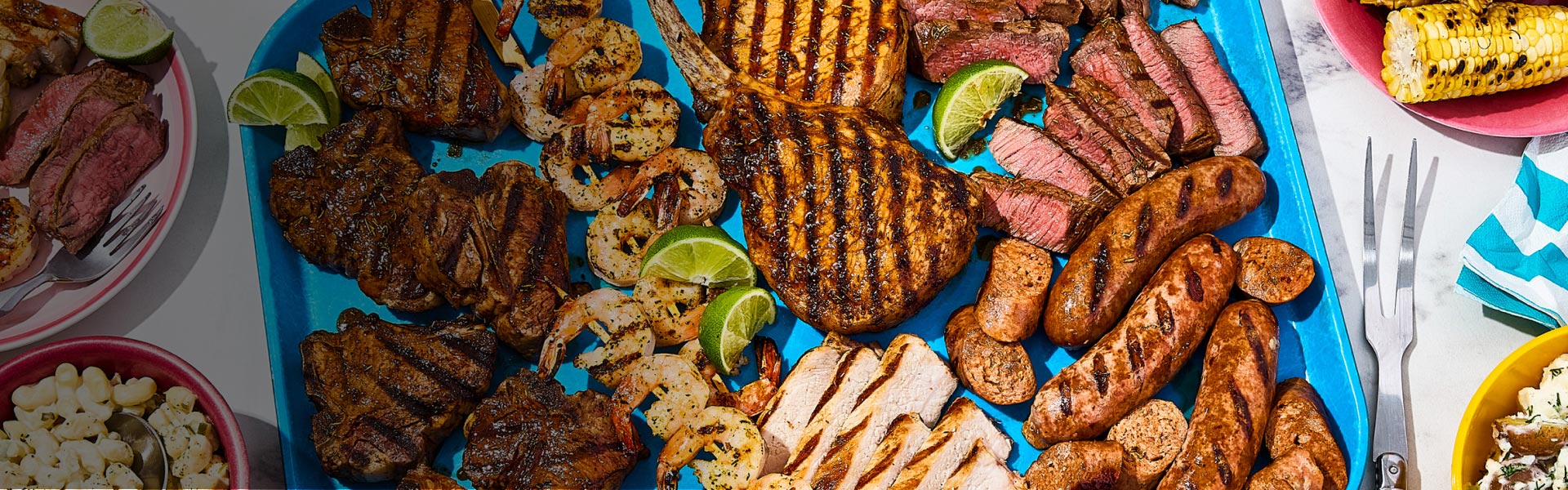 A large platter of all the Braai meats and seafood with a small plate of some off to the side, a small plate of grilled corn and two bowls with pasta and potatoes.