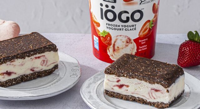Two plates with No-Bake Brownie Frozen Yogurt Sandwiches made with iÖGO Strawberry Swirl Frozen Yogurt. The dessert is surrounded by fresh strawberries, almonds in a small bowl, and a tub of frozen yogurt in the center.